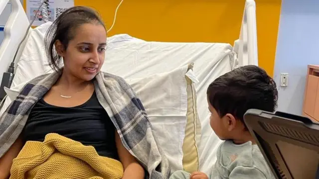 Sana en una cama de hospital junto a su hijo pequeño que la visita.