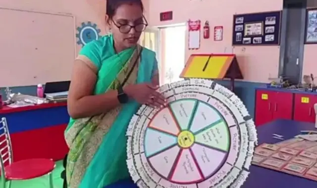 সবুজ শাড়ি, কালো ঘড়ি ও চোখে চশমা পড়া এক নারী একটি গোল চাকতির মতো একটি জিনিসি দেখাচ্ছেন যেটার মধ্যে ভাগ ভাগ করে কয়েকটি ঘরে কিছু লেখা আছে। পাশে চেয়ার-টেবিল দেখা যাচ্ছে 
