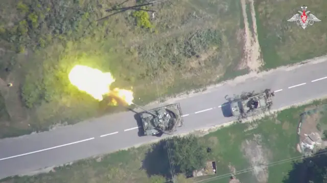 A imagem aérea mostra uma estrada com dois tanques de guerra. Um deles está atirando em direção ao lado esquerdo da foto. 