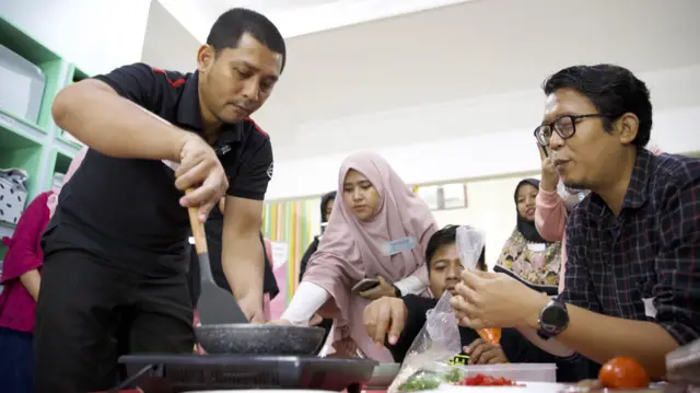 Rakean (kiri) dan seorang pria lain memasak di sekitar meja. Rakean sedang menggoreng telur dengan wajan di atas kompor, pria lainnya sedang membuka sekantong sayuran.