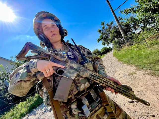 Oksana vestida com farda ucraniana e segurando fuzil enquanto sorri