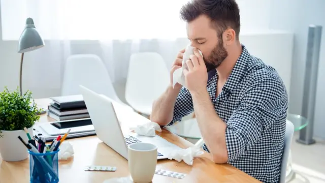 Un hombre en la oficina, resfriado, con pastillas y toallitas en el escritorio