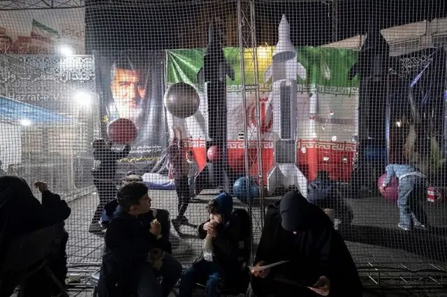 محوطه‌ای که با تور احاطه شده و کودکان در حال بازی هستند، و چند ماکت موشک، پرچم ایران و عکس یک فرمانده سپاه دیده می‌شود