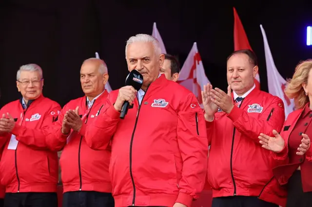 Binali Yıldırım ve TDT Aksakallar Konseyi üyeleri, üzerlerinde kırmızı Teknofest kıyafetleriyle bir sahnede. Yıldırım elinde bir mikrofonla konuşurken arkadakiler alkışlıyor.