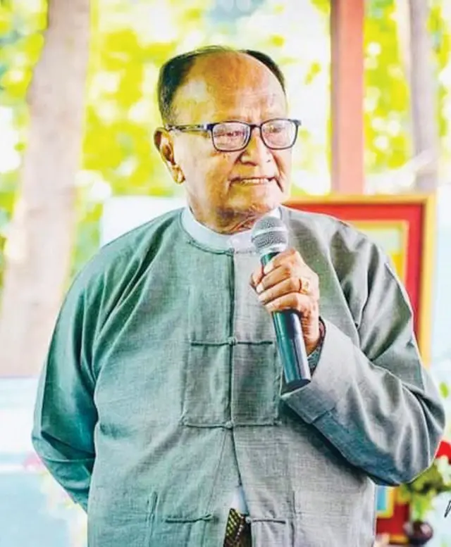 တိုက်ပုံအင်္ကျီခဲရောင်ဝတ်၊ မျက်မှန်တပ်ကာ မိုက်ကိုင် မတ်တပ်ရပ်စကားပြောနေတဲ့ ကဗျာဆရာကြီးကိုလေး