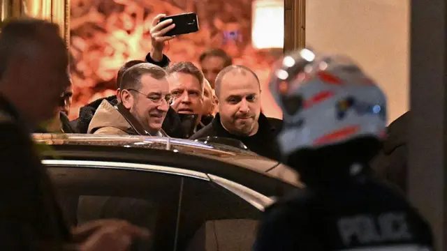 Умеров и еще несколько мужчин выходят из здания к автомобилю, на первом плане виден полицейский в шлеме