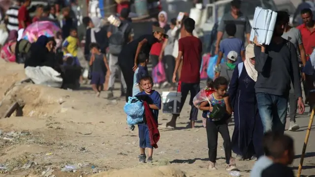 Un niño con una bolsa con ropa caminando junto decenas de personas más que caminan con sus pertenencias, otras llevan sus cosas en autos
