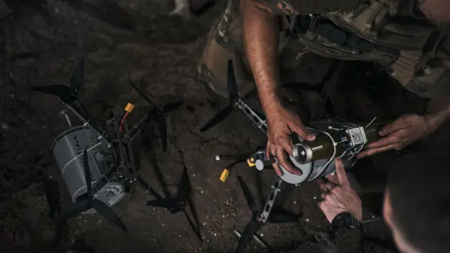 Un brazo sostiene un dron de color plateado posado en el suelo en la oscuridad.