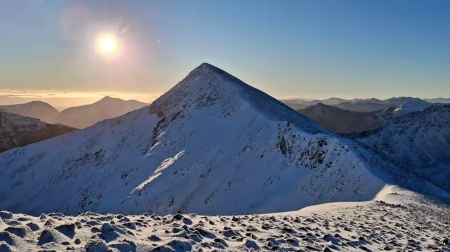 Un pico de montaña cubierto de nieve bajo un sol brillante, con crestas escarpadas y cordilleras distantes visibles contra un cielo azul claro.