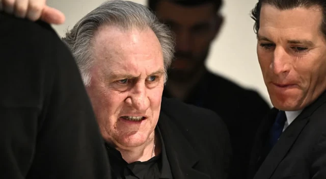 Depardieu zorlukla yürüyor ve yüzünü ekşitiyor. Yanında siyah takım elbiseli insanların arasında görünüyor.