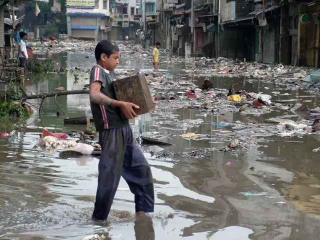 An Indian boy carries his belonging through a flooded street strewn debris in Surat, in the western state of Gujurat, 13 August 2006. Floods have killed at least 65 people in the Indian diamond-cutting city of Surat, officials said 12 August, as slowly receding waters allowed city officials to recover corpses. 
