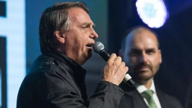 Sorrindo, Eduardo Bolsonaro observa seu pai discursando