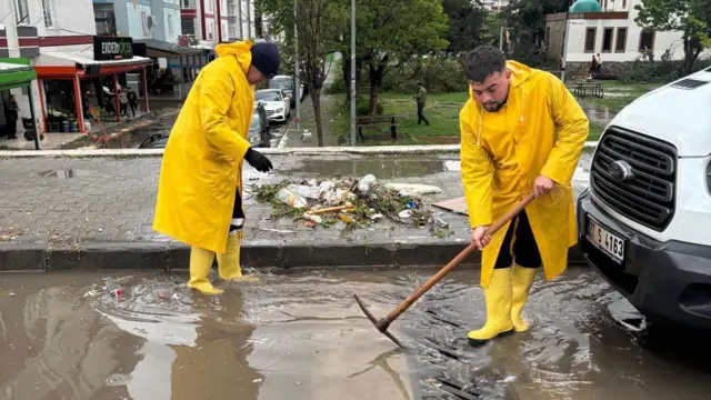 Sarı yağmurluklu ve çizmeli belediye çalışanları yol kenarındaki bir gideri açmaya çalışıyor. Çalışanlardan birinin elinde kazma var.