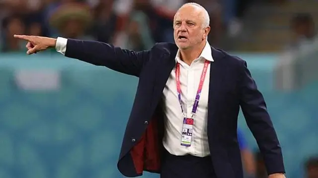 غراهام أرنولد بلباس رسمي أسود وقميص أبيض دون ربطة عنق، يؤشر بيده ويتحدث.