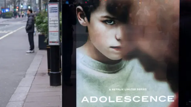 A poster for the film Adolescence on a bus stop. It shows a young boy wearing a grey sweatshirt looking at his his father, who is out of focus.