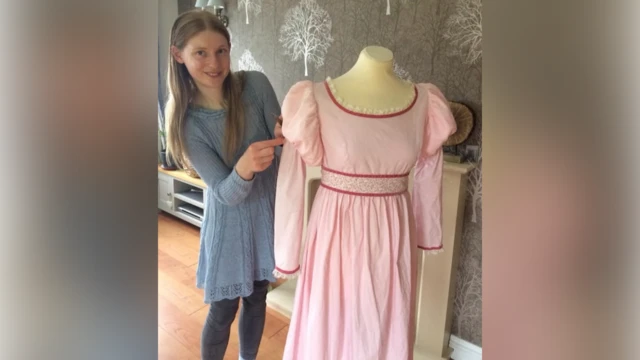 Una joven de cabello castaño largo con un vestido azul claro está de pie junto a un maniquí. Este lleva un vestido rosa de mangas largas y hombros abullonados. Lleva un cinturón en la cintura.