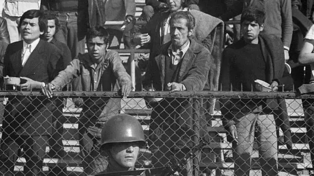 Photo en noir et blanc montrant des personnes détenues dans un stade de sport à la suite du coup d'État. Un soldat armé d'un fusil se tient devant la clôture, les prisonniers attendent leur tour pour être interrogés.