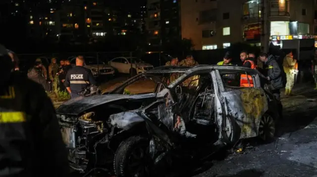  夜の街で、黒焦げになった車両の周りに「POLICE」と書かれたジャケットを着た男性や蛍光色のベストを着た男性たちが立っている