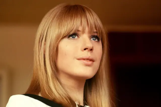 Marianne Faithfull pictured in the 1960s. She has straight, shoulder-length light brown hair and blunt bangs, and is wearing a white top with a black collar detail. The background appears to be indoors with warm lighting and a dark area behind, suggesting an interior setting.