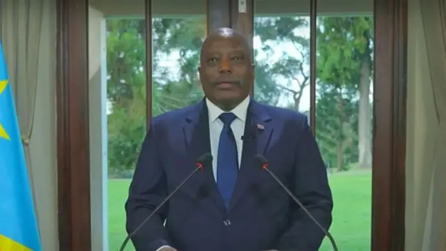 Joseph Kabila ahagaze imbere y'indangururamajwi ebyiri arimo kuvuga ijambo. Yambaye ikositimu y'ubururu na karuvati, n'ishati y'umweru, iruhande rwe hari ibendera rya DR Congo.