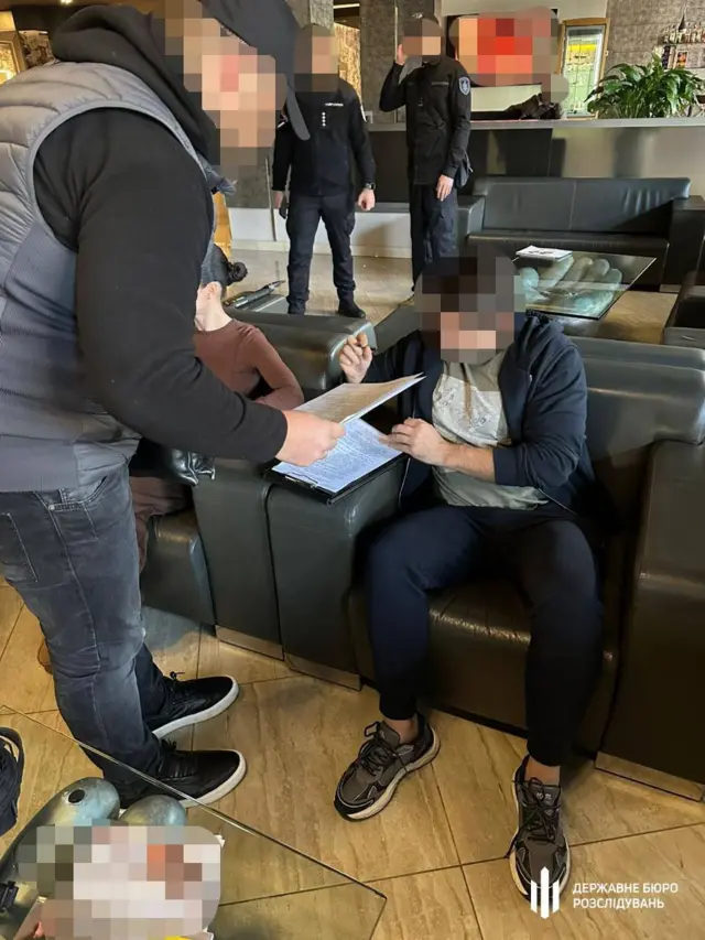 Затриманий із заблюреним обличчям підписує документи в готельному лоббі, позаду стоять двоє працівників поліції