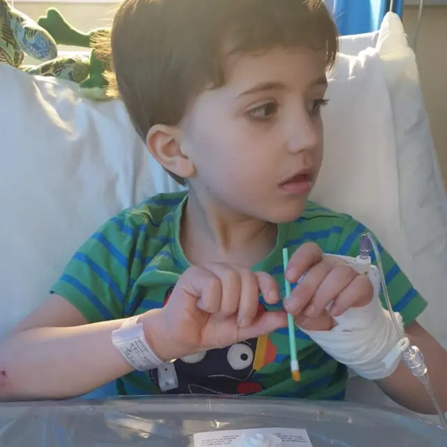 Criança sentada na cama, com uma mão enfaixada e um tubo médico, segurando um pequeno swab