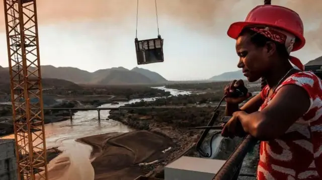 یو کارګر چې سور رنګ خولۍ یې پر سر ده او د لمر خته پر مهال د بند د جوړولو ځای ته ګوري. یو کرین هم لېدل چې کېدای شي، د کانکرېټ یوه لویه تخته پورته کوي.