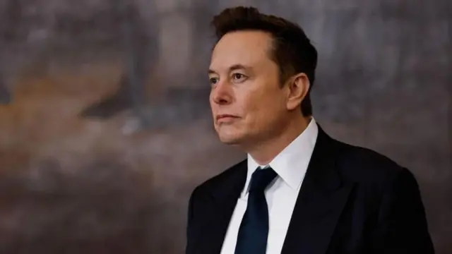 Elon Musk vestindo um terno preto, camisa branca e gravata preta olhando para longe