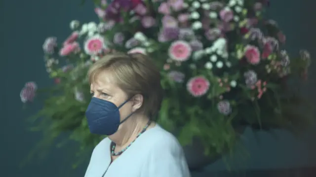 Kanclerz Niemiec Angela Merkel widoczna przez szybę, w maseczce FFP2, stoi przed obrazem przedstawiającym kwiaty w Urzędzie Kanclerskim w Berlinie, w czerwcu 2021 roku.