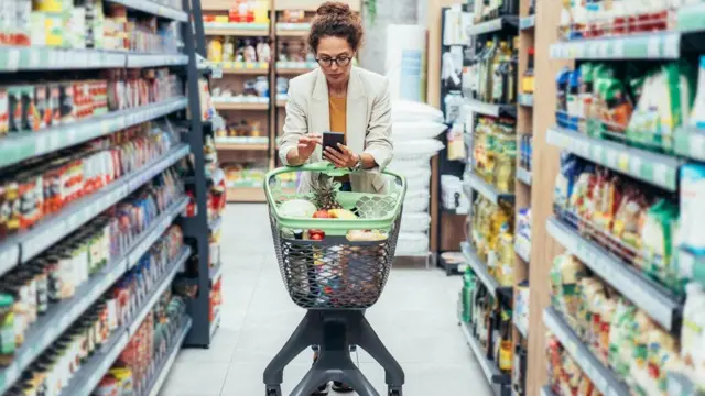 Mulher anda com carrinho de compras em supermercado enquanto mexe no celular