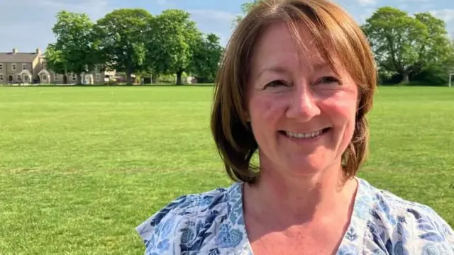 Catherine Judkins está de pie en un campo deportivo. La foto muestra su cabeza y hombros, y se puede ver la portería de fútbol al fondo, a un lado, y una casa que bordea el campo, al otro. Catherine lleva una blusa de manga corta con cuello en V y estampado floral azul y blanco. Catherine sonríe a la cámara.