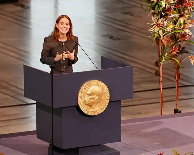 Женщина в коричневом костюме стоит на трибуне с профилем Альфреда Нобеля