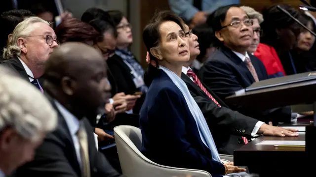 11 डिसेंबर 2019 ला हेगमधील पीस पॅलेसमध्ये नरसंहाराच्या आरोपासंदर्भातील सुनावणीच्या दुसऱ्या दिवशी, न्यायालयात गॅम्बियाचे न्यायमंत्री (डावीकडून दुसरे) अबुबकर तांबाडू यांच्याजवळ बसलेल्या आंग सान सू की. त्यांच्या आजूबाजूच्या लोकांबरोबर त्या वरच्या दिशेनं पाहत आहेत. त्यांनी गडद रंगाचं सूट जॅकेट आणि फिकट निळ्या रंगाचा ब्लाऊज घातला आहे.