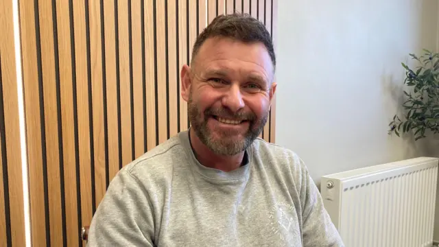 Un hombre sonriente, de pelo corto y oscuro, y barba, está sentado en el interior. Lleva una sudadera gris claro y está sentado frente a unos paneles de madera en la pared.