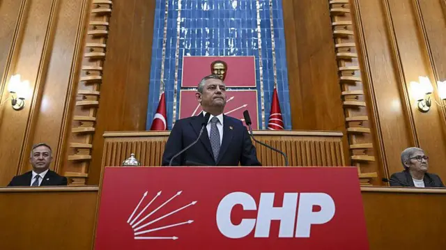 CHP lideri Özgür Özel meclisteki grup toplantısında kürsüde konuşuyor, arkasında Atatürk büstü ve bayraklar, iki yanında oturan bir erkek bir kadın iki üye var