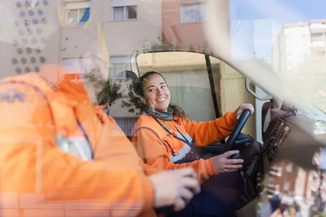 La conductora de una ambulancia, con una gran chaqueta naranja, sonríe a su compañero quien está sentado en el asiento del copiloto.