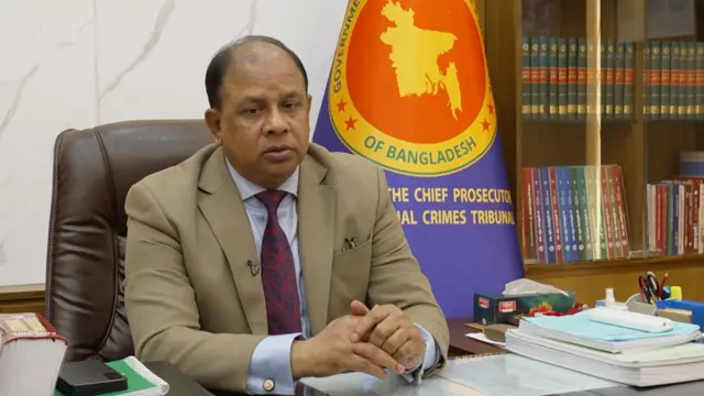 আন্তর্জাতিক অপরাধ ট্রাইব্যুনালের চিফ প্রসিকিউটর মো.আমিনুল ইসলাম বাদামী কোট পরে বসে আছেন। তিনি দুই হাত কাচের টেবিলের ওপর তুলে রেখেছেন। পেছনে নীলের মাঝখানে হলুদ বর্ডারের মাঝে লাল বৃত্তের ভেতরে বাংলাদেশের মানচিত্র।  ইংরেজিতে আন্তর্জাতিক অপরাধ ট্রাইব্যুনাল ও  চিফ প্রসিকিউটর লেখা। পেছনে বুক শেলফে কিছু বই। 