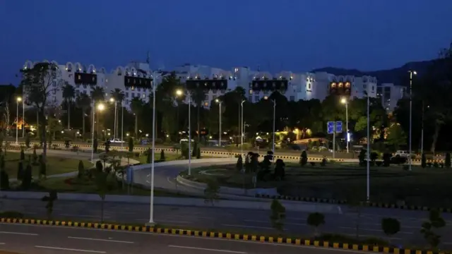 夜の空の下、多くの街灯がついた道路の向こうに、白い複数階の建物が横に大きく広がっている。建物内の照明もたくさんついている