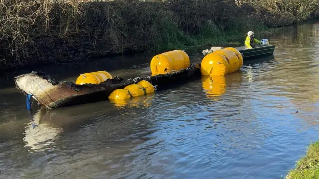 Un barco abandonado siendo remolcado con bolsas inflables especiales en un canal.