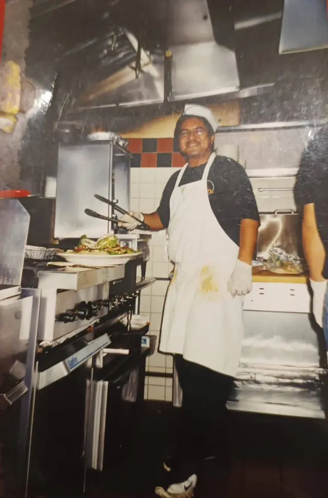 Hombre con delantal trabajando en una cocina industrial, usando pinzas frente a una parrilla con comida, rodeado de equipos de metal.
