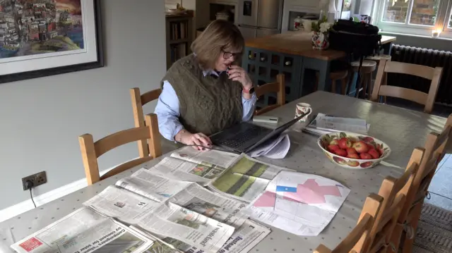 Una mujer con gafas y cabello oscuro, vestida con un jersey de punto marrón, está sentada en una mesa de cocina con un portátil y periódicos delante. Hay un bol de manzanas rojas sobre la mesa y un mapa tamaño A3 con zonas en rosa, posiblemente la ubicación de un parque solar.