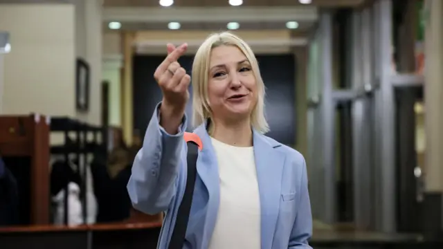 Екатерина Дунцова, светловолосая молодая женщина в голубом пиджаке, улыбается и показывает пальцами знак "сердце"