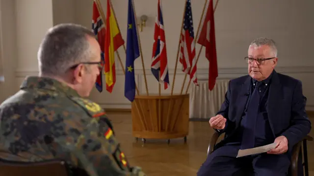 El general Carsten Breuer en sus fatigas militares sentado frenta a  Allan Little, vestido con un traje oscuro.