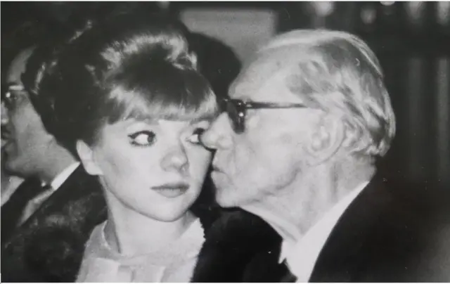 Una imagen cerrada en blanco y negro de una joven mujer con un peinado elegante y un hombre mayor de perfil con anteojos