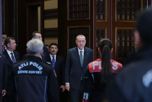 Cumhurbaşkanı Erdoğan kameralara doğru yürüyor, önünde polis üniforması giymiş kişiler var.