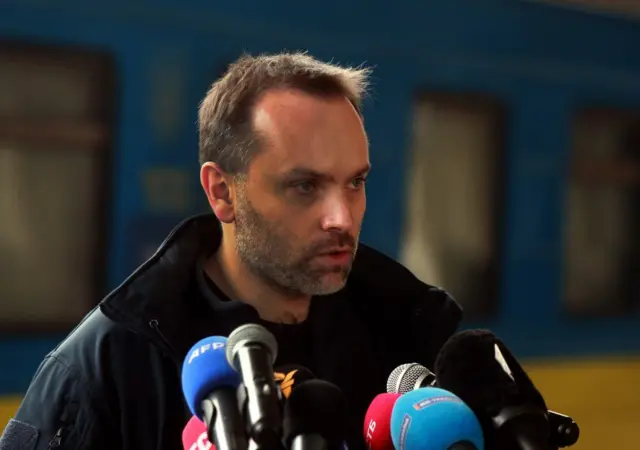 Олександр Перцовський у темній куртці стоїть перед мікрофонами різного кольору