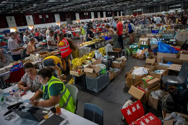 Un enorme centro de refugio para las personas evacuadas. Se ven muchísimas cajas con insumos básicos, como mantas protectoras, comida y demás. Hay muchas personas voluntarias que visten chalecos reflectantes organizando el lugar y entregando insumos. También se ve mucha gente haciendo cola para recoger cosas o esperando al fondo. 