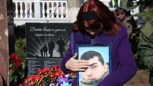 Жінка тримає портрет молодого чоловіка в уніформі. Вона вдягнена у синє пальто, на голові - чорна траурна пов'язка. Позаду - чорний мраморний пам'ятник загиблим у війні.