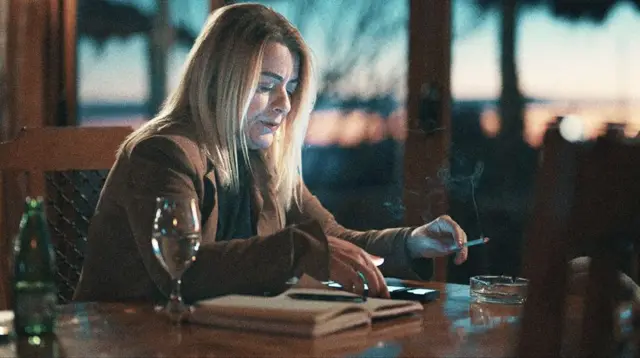 Гордана Вуйсич — женщина с длинными светлыми волосами в коричневом пиджаке сидит за деревянным столом у окна с видом на закат. В ее руке сигарета, она внимательно изучает что-то на экране телефона, рядом с ней блокнот, бокал и пепельница.