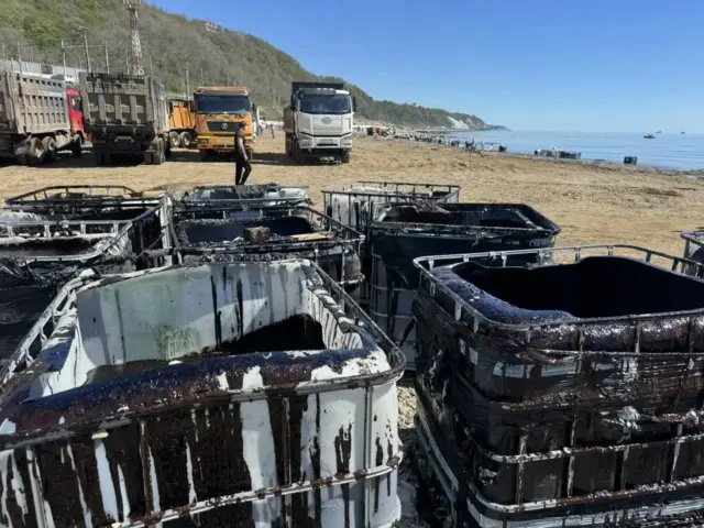 На переднем плане видны бочки для сбора нефти, установленные на пляжах. За ними можно увидеть море и край берега 